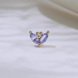Peut inclure: Une petite boucle d'oreille dorée avec un motif en forme de croissant. La boucle d'oreille présente deux pierres précieuses de couleur violet clair, de taille marquise, et trois petites perles dorées. Le bijou est posé sur un fond blanc.