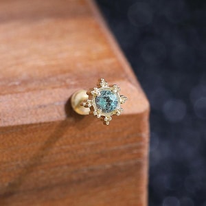 Puede incluir: Un pendiente de botón de oro con una piedra preciosa redonda de color azul verdoso en el centro, rodeada por un engaste de metal en forma de estrella con pequeñas cuentas decorativas en cada punta. Un cierre redondo de oro es visible.