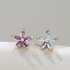 Op de afbeelding: Twee oorknopjes in de vorm van vijfbladige bloemen. De ene oorbel is voorzien van paarse edelstenen in marquise-slijpvorm, de andere van lichtblauwe edelstenen in marquise-slijpvorm. Beide zijn gezet in goudkleurig metaal met een gouden kraal.