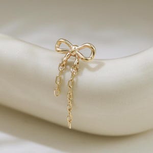 Puede incluir: Un pendiente de tono dorado que presenta una pequeña y delicada forma de lazo en la parte superior. Dos cadenas finas de eslabones cuelgan verticalmente desde el centro del lazo, creando un efecto colgante. El metal tiene un acabado pulido.