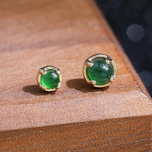 May include: A pair of stud earrings featuring small, round, cabochon-cut green stones set in gold-toned metal prongs. The earrings are displayed side-by-side, showcasing the smooth, polished surface of the green gems and the metallic setting.