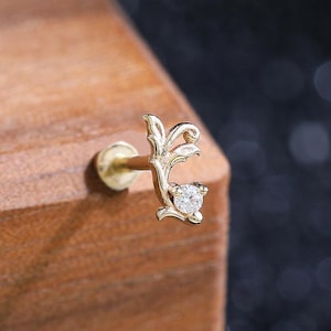 May include: A gold stud earring featuring a delicate, curved vine-like design with a single small, clear round gemstone set at the tip. The metal has a polished finish, and the post is visible extending from the back of the decorative element.