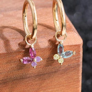 Puede incluir: Un par de pendientes de aro dorados, cada uno con un pequeño dije en forma de mariposa. Una mariposa está compuesta por cuatro piedras preciosas rosas de talla marquesa, mientras que la otra tiene dos piedras azules y dos amarillas de talla marquesa.