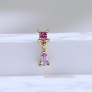 Könnte beinhalten: Goldfarbener Ohrring mit einem quadratischen, magentafarbenen Edelstein oben und einem tropfenförmigen, hellvioletten Edelstein, der darunter hängt. Der Ohrring ist vor einem weißen Hintergrund mit weicher Beleuchtung platziert, die das Design hervorhebt.