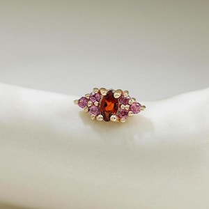 Può includere: Un piccolo orecchino a lobo in oro con una pietra preziosa centrale di colore rosso intenso a forma ovale, affiancata da grappoli di minuscole pietre rotonde rosa su entrambi i lati. Il gioiello presenta un design delicato e simmetrico.