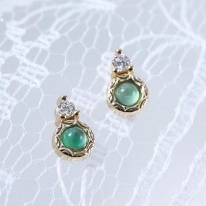 May include: A pair of gold stud earrings, each featuring a round, cabochon-cut green stone set within a decorative gold frame, topped with a single, small, clear, round-cut gemstone. The earrings are displayed on a white lace fabric.