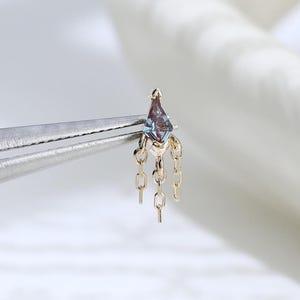 Puede incluir: Un pequeño pendiente de oro con una piedra preciosa azulada en forma de cometa en la parte superior y tres delicadas cadenas doradas colgantes. La pieza está sujeta por unas pinzas de metal plateado, mostrando su diseño detallado.