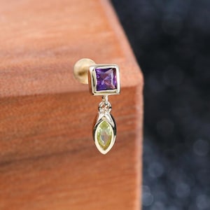 May include: A gold stud earring featuring a square-cut purple gemstone at the top and a dangling marquise-cut green gemstone below. The earring has a post back and is constructed from a metallic material with a polished finish, showcasing two distinct gemstone shapes and colors.