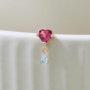 May include: A gold-colored earring featuring a heart-shaped, deep pink gemstone at the top. Below the heart is a small, light blue, teardrop-shaped gemstone. The earring is set against a white background.