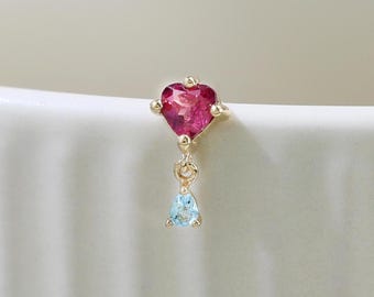 May include: A gold-colored earring featuring a heart-shaped, deep pink gemstone at the top. Below the heart is a small, light blue, teardrop-shaped gemstone. The earring is set against a white background.