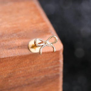 May include: A small gold-toned stud earring featuring a delicate bow design. The earring has a flat circular backing and a thin post, with the bow element positioned on the front. The metal has a polished finish that reflects light.