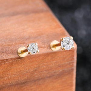 Puede incluir: Dos pendientes de botón de oro, cada uno con una piedra preciosa redonda, transparente y facetada en un engaste de cuatro garras. Los pendientes tienen un cierre plano y circular y están colocados sobre una superficie de madera clara.