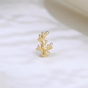 May include: A small, gold-colored stud earring in the shape of a coral branch. The earring is centered on a white marble surface, with a blurred, off-white background. The earring is a delicate piece of jewelry.