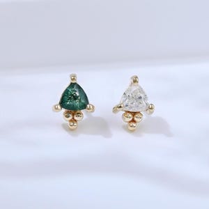 May include: A pair of gold stud earrings. One earring features a triangular, emerald-green gemstone, while the other has a clear, triangular gemstone. Both earrings have a cluster of small gold beads at the base.