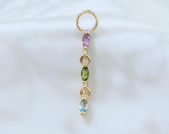 May include: A gold-colored earring with a small hoop and a dangling chain of colorful gemstones. The gemstones are oval-shaped and include purple, green, and blue hues. The earring is displayed against a white, textured background.