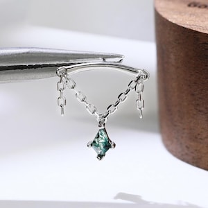 Puede incluir: Una joya corporal de plata con una barra curva y dos delicadas cadenas colgantes que se unen en un punto central, suspendiendo una pequeña piedra preciosa de ágata musgosa verde en forma de cometa. El metal es plata pulida.