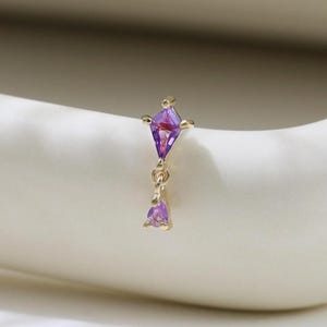 May include: A gold-colored nose ring featuring a purple kite-shaped gem at the top and a dangling purple teardrop-shaped gem below. The jewelry is set against a white background.