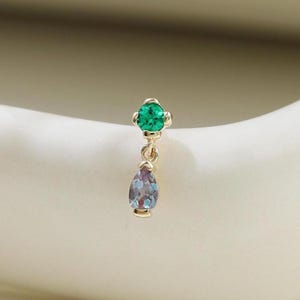 May include: A single gold earring with two gemstones. The top gemstone is a round, bright green color. The bottom gemstone is a teardrop shape with a purple and blue color. The earring is set against a white background.
