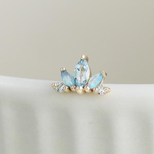 May include: A gold-toned stud earring featuring a central marquise-cut light blue stone flanked by two smaller marquise-cut light blue stones and two tiny clear round stones, arranged in a symmetrical, fan-like cluster design on a metal base.