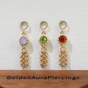 Peut inclure: Trois boucles d'oreilles dorées avec des pierres précieuses colorées : violet, vert et rouge. Chaque boucle d'oreille a un motif de chaîne. Elles sont présentées sur un présentoir en bois avec le texte "Golden Aura Piercings".