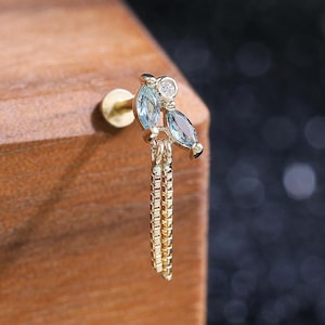 May include: A gold stud earring featuring two marquise-cut light blue stones and one small round clear stone arranged in a cluster, with two delicate gold chains hanging vertically from the base of the setting, secured by a flat gold back.