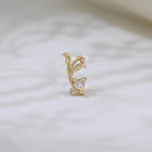 May include: A gold-colored earring with an ornate, leaf-like design. The earring features a small, clear gemstone at the bottom. The earring is set against a white marble surface.