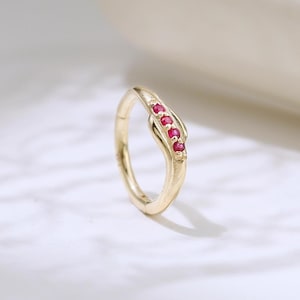 May include: A gold-colored ring with a curved design, featuring a series of small, vibrant red gemstones. The ring is set against a soft, neutral background, highlighting its elegant and delicate features. This piece of jewelry is suitable for everyday wear or special occasions.