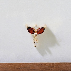 May include: A gold-coloured piercing featuring red and white gemstones. The design includes two red, marquise-cut stones and smaller white stones. The text "Golden Aura Piercings" is visible on a wooden surface. A unique piece of jewellery.