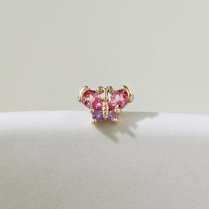 Puede incluir: Un pendiente de botón con forma de mariposa de color dorado. Las alas de la mariposa están adornadas con piedras preciosas rosas y moradas. El pendiente se coloca sobre un fondo neutro, resaltando su delicado diseño.