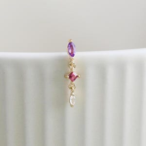 May include: A gold earring with a vertical design. It features an elongated purple gemstone at the top, followed by a small red gemstone, a tiny white gemstone, and a dangling clear gemstone. The earring is set against a white background.