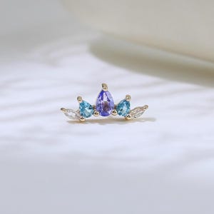Puede incluir: Una pequeña joya dorada en forma de corona. La corona presenta una gema central morada en forma de lágrima, flanqueada por dos gemas azules y gemas blancas más pequeñas. Sobre un fondo blanco suave.
