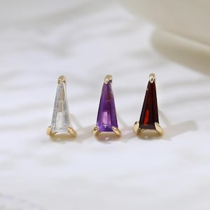 May include: Three gold stud earrings with triangular gemstone accents. The gemstones are clear, purple, and red. The earrings are displayed on a white, marble-effect surface.