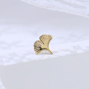 May include: A small gold stud earring shaped like a ginkgo leaf. The metal piece features a polished finish with delicate, radiating engraved lines that mimic the natural veins of a leaf, tapering to a point at the base of the stem.