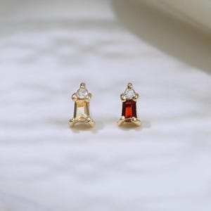 May include: Two gold-colored stud earrings. One earring features a clear, round gemstone, and the other has a rectangular, red gemstone. The earrings are set on a white, textured surface.
