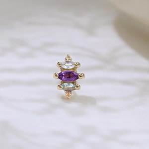 Könnte beinhalten: Ein goldfarbener Ohrstecker mit drei Edelsteinen: einem zentralen, marquiseförmigen, lila Amethysten, flankiert von zwei kleineren, klaren, oval geschliffenen Steinen. Der Ohrring hat ein filigranes Design, für Ohrlöcher.