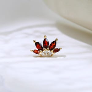 May include: A gold-coloured stud earring with a crown design. The crown features five marquise-cut, deep red gemstones and several small, round, clear gemstones. The earring is set against a white background with a curved, white ceramic object.