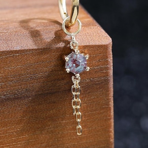 Puede incluir: Un pendiente de aro dorado con un pequeño dije de piedra preciosa hexagonal iridiscente suspendido de una delicada cadena de oro. El pendiente está hecho de oro amarillo pulido, con la piedra mostrando sutiles tonos azules y morados.