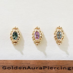 May include: Three gold earrings with oval gemstones in green, purple, and blue. Each earring has a decorative, filigree border. The earrings are displayed on a wooden stand with the text "Golden Aura Piercings".