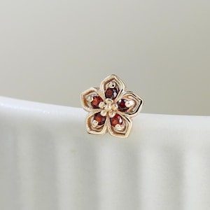 May include: A small gold stud earring shaped like a five-petaled flower. Each petal features a deep red gemstone set within a polished gold frame, with a tiny gold bead at the center of the flower, creating a delicate and detailed floral design.