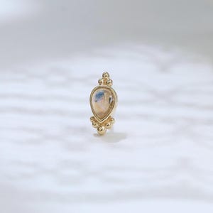May include: A gold-colored earring with a teardrop-shaped, light-colored stone. The earring has a decorative design with small, round accents at the top and bottom. The stone has a subtle, multi-toned appearance.