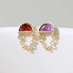 May include: A pair of gold earrings. Each earring has a half-circle gemstone, one red and one purple, hanging from a chain. The earrings are presented on a white surface.