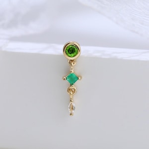 May include: A gold earring featuring a green gemstone, a green square-shaped gemstone, and a small clear gemstone. The earring is displayed against a white backdrop with a delicate lace detail. The design is elegant.