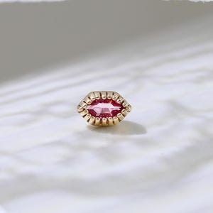 May include: A gold-colored stud earring with a pink, marquise-cut gemstone. The earring is surrounded by a beaded border. The background is a soft white, with a lace detail in the upper left corner.