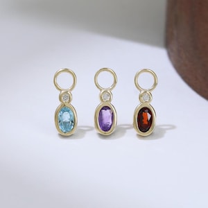 Pode incluir: Três pingentes dourados com pedras preciosas ovais azuis, roxas e vermelhas. Cada pingente tem um pequeno detalhe de diamante e um laço circular no topo para pendurar. Os pingentes são exibidos em uma superfície branca. As pedras medem cerca de 1 cm.