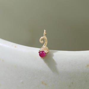 May include: A small, gold-colored pendant with a swirling design and a round, red gemstone at the base. The pendant is set against a soft, off-white background, highlighting its delicate features and vibrant color.