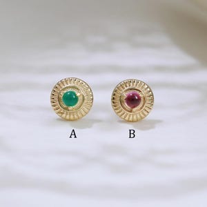 以下が含まれることがあります： 中央に宝石が付いた2つのゴールドスタッドピアス。片方のピアスは緑色の宝石、もう片方はピンク色の宝石です。ピアスにはAとBのラベルが付いています。白い表面にあります。