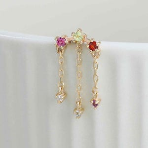 May include: A gold earring featuring a curved bar set with three small, colourful gemstones in pink, light green, and red. Three delicate gold chains hang from the bar, each ending in a tiny, clear, sparkling gemstone accent.