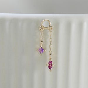 Könnte beinhalten: Ein goldener Schmuckanhänger mit einem kleinen runden Verbindungsring, an dem zwei zarte Goldketten unterschiedlicher Länge befestigt sind. Eine Kette endet in einem quadratisch geschliffenen violetten Edelstein, die längere in einem pinken Markise-Edelstein.