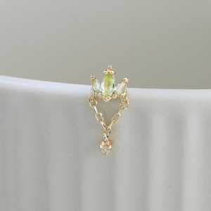 May include: A gold stud earring featuring a marquise-cut light green gemstone at the top, connected by two delicate gold chains to a small, round, clear gemstone dangling at the bottom, creating a tiered, dangling design.