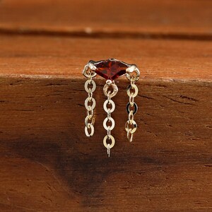May include: A gold piercing jewellery piece featuring a marquise-cut deep red gemstone at the top, with three delicate gold chains hanging down from the setting. The text GoldenAuraPiercings is printed in black on the wooden display block.